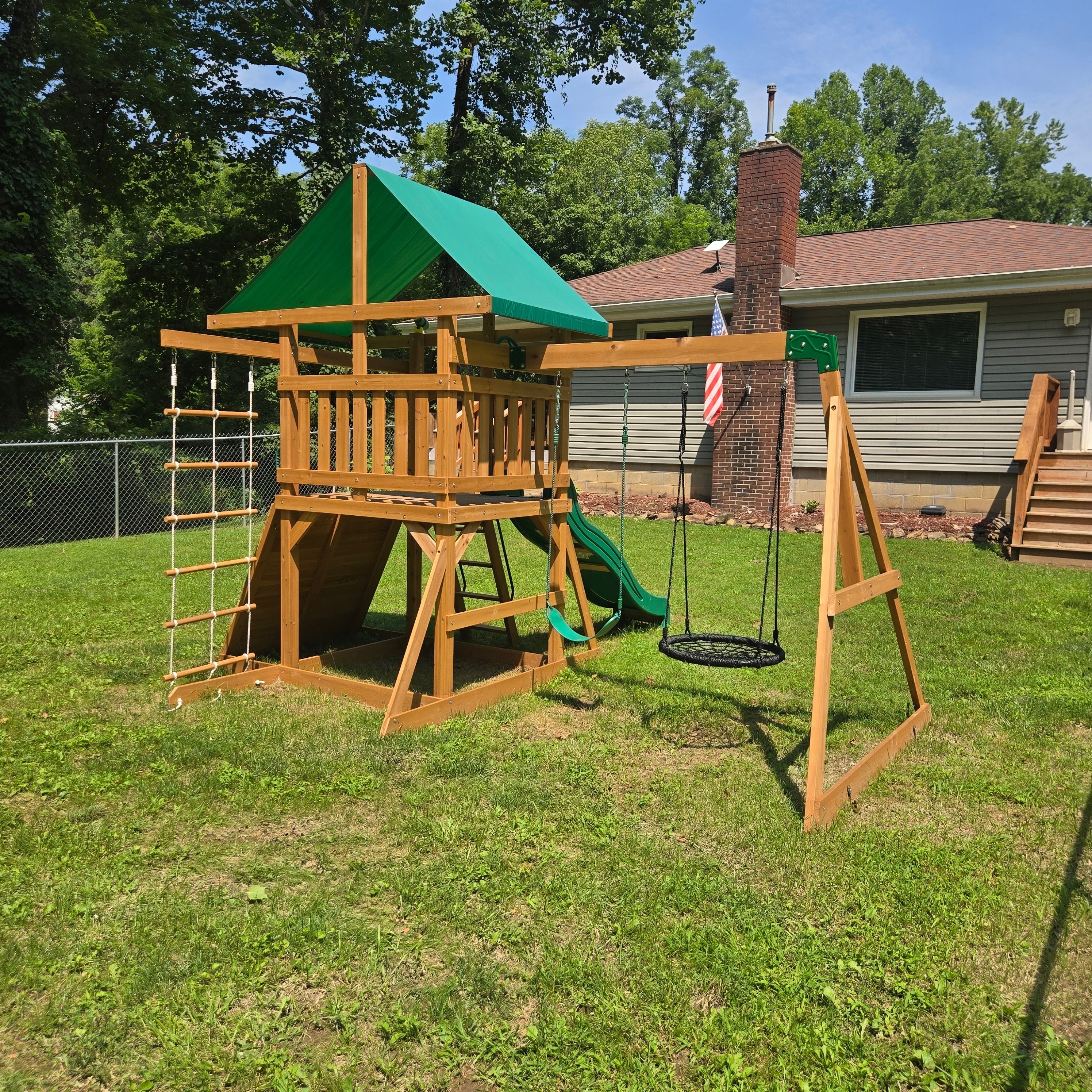 Wooden playset with slide, swings, climbing rope ladder and sandbox in the fenced backyard